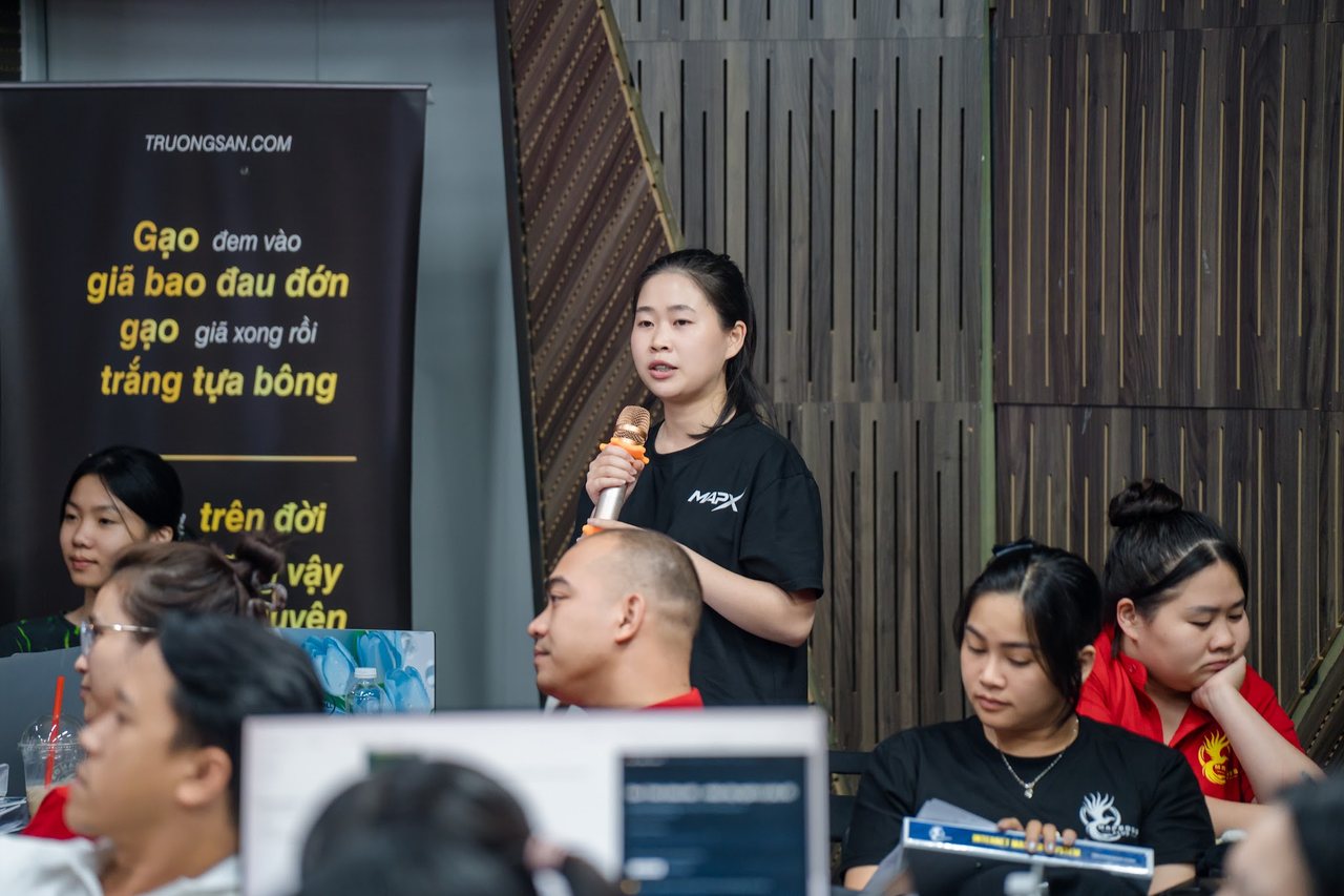 Học viên IMS mùa 2 cầm micro chia sẻ cạnh banner câu thơ hạt gạo