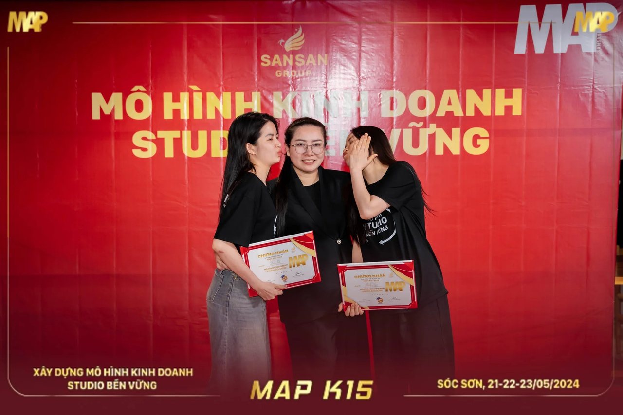 Hai học viên MAP CAMP mùa 15 hôn má người đứng giữa trước phông đỏ
