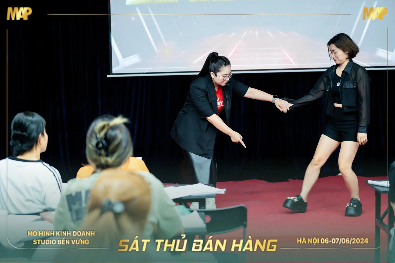 Người dẫn dắt Sát Thủ Bán Hàng nắm tay học viên làm mẫu trên sân khấu