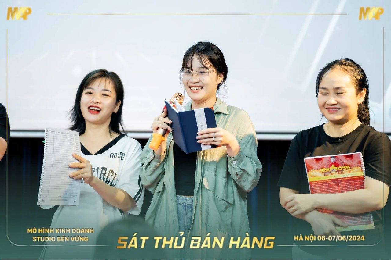 Nhóm học viên Sát Thủ Bán Hàng cầm micro và tài liệu cười trên sân khấu