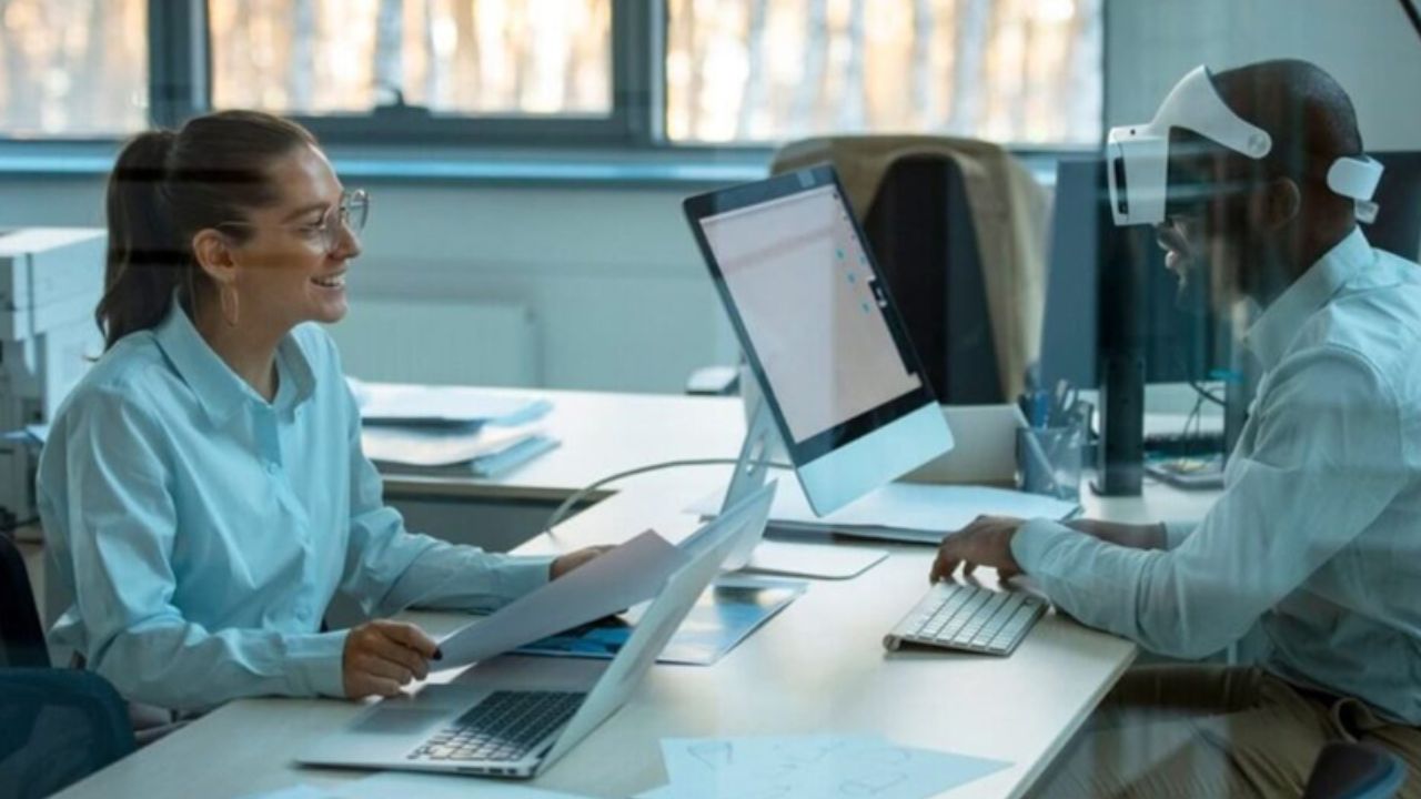 Hai nhân viên văn phòng trao đổi công việc tại bàn làm việc có máy tính và tài liệu, minh họa những vị trí hành chính, xử lý hồ sơ và tuyển dụng đang chịu tác động từ trí tuệ nhân tạo (AI).