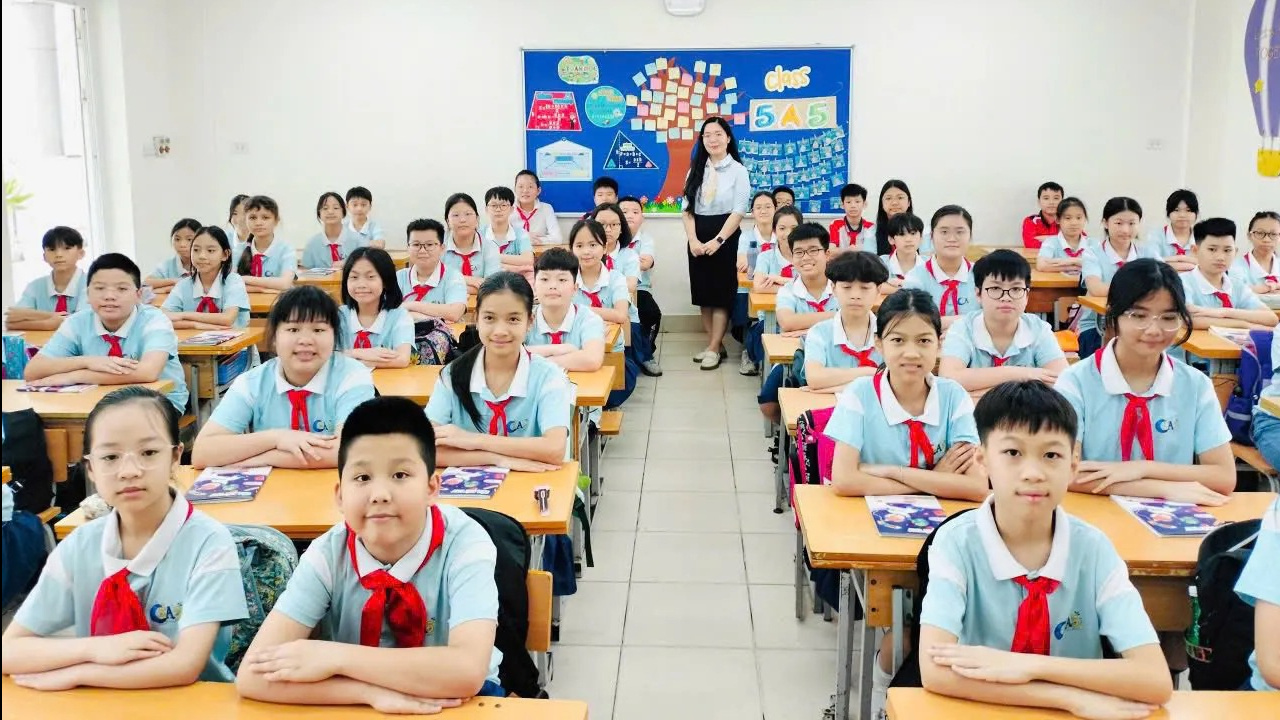 Lớp học tại Việt Nam với đông học sinh mặc đồng phục, đeo khăn quàng đỏ ngồi ngay ngắn tại bàn học, giáo viên đứng phía cuối lớp, minh họa môi trường giáo dục và đầu tư học tập cho con.